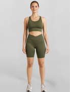 Woman wearing an olive green khaki bike shorts and sports bra athletic outfit on a gray background